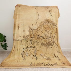 May include: A large, tan throw blanket featuring a detailed map design. The map is in shades of brown and black, with intricate illustrations of mountains, forests, and bodies of water. The blanket is draped over a chair.