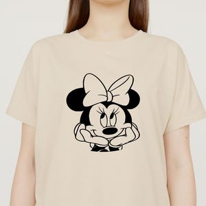 Peut inclure: Un t-shirt beige avec une illustration en noir et blanc de Minnie Mouse. Minnie Mouse porte un n&oelig;ud et a une expression espi&egrave;gle.