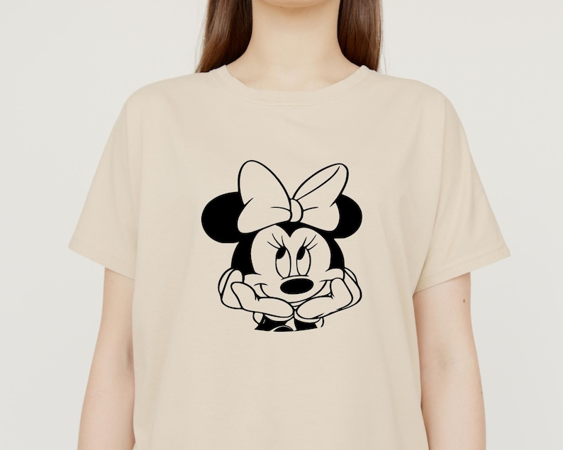 Peut inclure: Un t-shirt beige avec une illustration en noir et blanc de Minnie Mouse. Minnie Mouse porte un n&oelig;ud et a une expression espi&egrave;gle.
