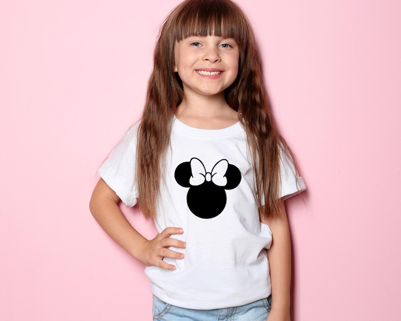 Peut inclure: Un t-shirt blanc avec une image noire et blanche de la t&ecirc;te de Minnie Mouse avec un n&oelig;ud sur le dessus.