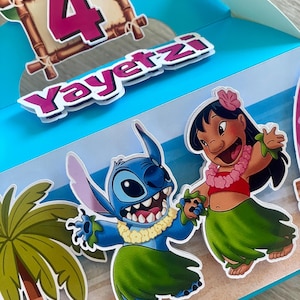 May include: A blue party favor box featuring the characters Stitch and Lilo from the movie Lilo & Stitch. The box has a tropical theme with palm trees, a sandy beach, and the number 4. The name "Yayertzi" is also displayed.