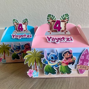 May include: Two colorful party favor boxes, one blue and one pink, feature a tropical beach scene with cartoon characters. Each box has the number "4" and the name "Yayetz" on top. The boxes are decorated with palm trees and a surfboard.