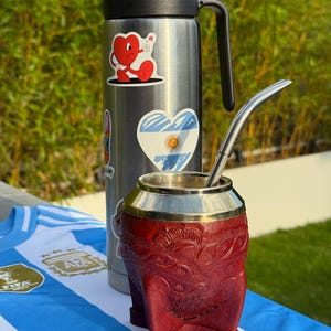 Peut inclure: Une gourde isotherme en acier inoxydable avec une poignée noire et des autocollants, à côté d'une tasse à maté en cuir rouge avec un bord en métal et une paille. Un maillot de football argentin est en arrière-plan.