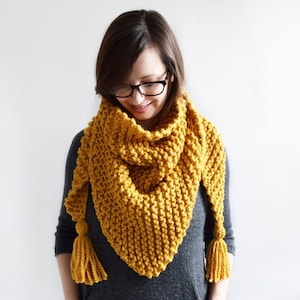 May include: A mustard yellow knitted scarf with tassels. The scarf is triangular in shape and is worn by a person with brown hair and glasses.
