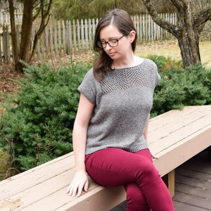 Puede incluir: Una mujer con un top de punto gris con un escote de encaje y pantalones marrones est&aacute; sentada en un banco de madera. Lleva sandalias marrones.
