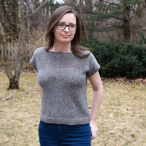 Puede incluir: Una mujer lleva una blusa de punto gris con un escote de encaje y unos vaqueros azules. La blusa tiene mangas cortas y un corte holgado.