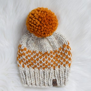 May include: A knitted beanie with a pom pom on top. The beanie is white and mustard yellow with a striped pattern. The beanie has a brown leather tag with the letters "KK" on it.