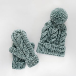 May include: A set of teal blue knitted winter accessories including a hat with a pom pom and matching mittens.