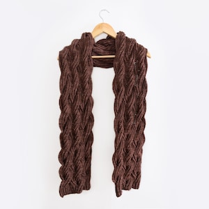 Pode incluir: Uma echarpe de malha castanho com um padrão de trança, pendurada num cabide de madeira sobre um fundo branco.
