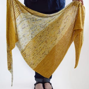 May include: A knitted shawl in shades of yellow, white, and gray. The shawl features a striped pattern with a gradient effect, transitioning from a lighter hue to a deeper yellow. The fabric appears soft and lightweight.