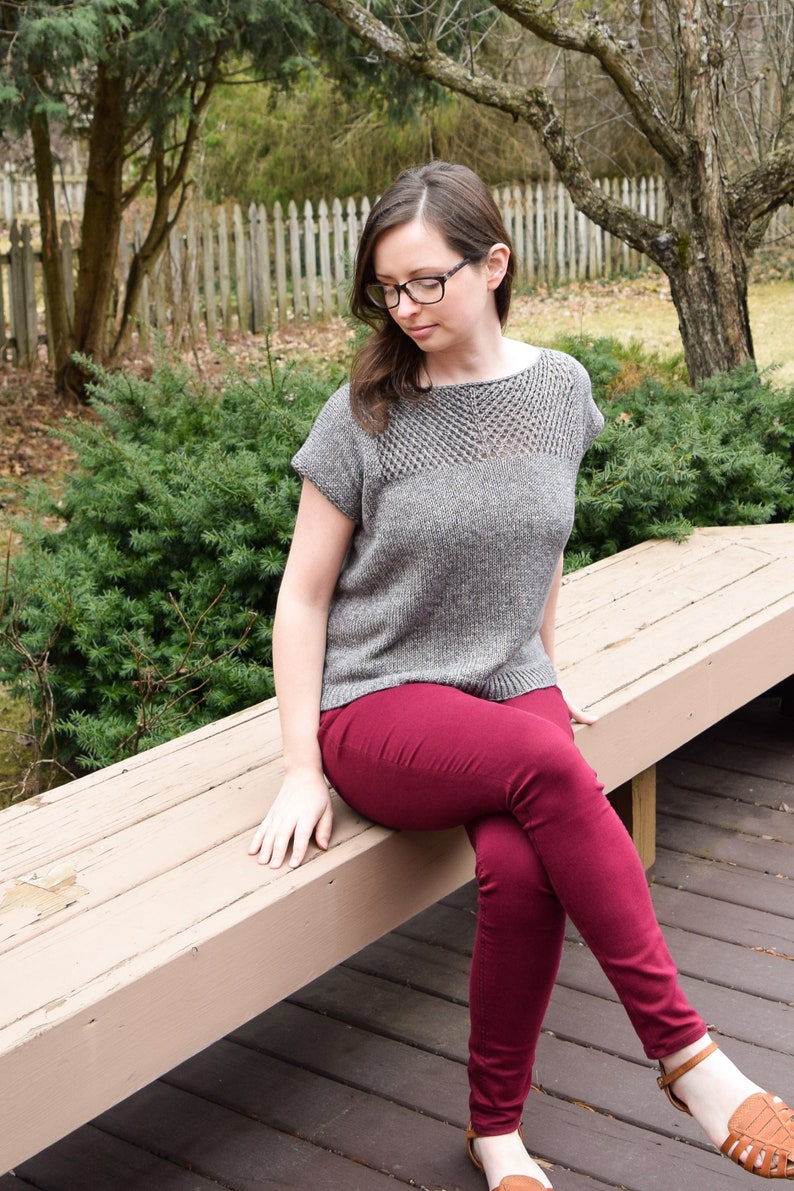 Puede incluir: Una mujer con un top de punto gris con un escote de encaje y pantalones marrones est&aacute; sentada en un banco de madera. Lleva sandalias marrones.