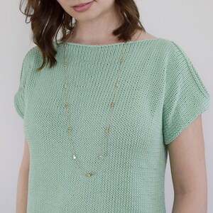 May include: A light green knitted top with short sleeves and a scoop neck. The top features a gold chain necklace with small square-shaped charms.