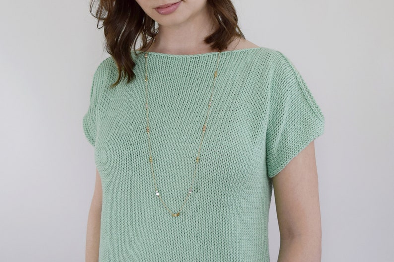 May include: A light green knitted top with short sleeves and a scoop neck. The top features a gold chain necklace with small square-shaped charms.