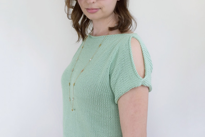 May include: A light green knitted top with a scoop neck and short sleeves. The sleeves have cutouts at the shoulders. The top is worn with a gold necklace.