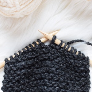 A work in progress photo of a charcoal colored scarf with cream colored yarn in the background.