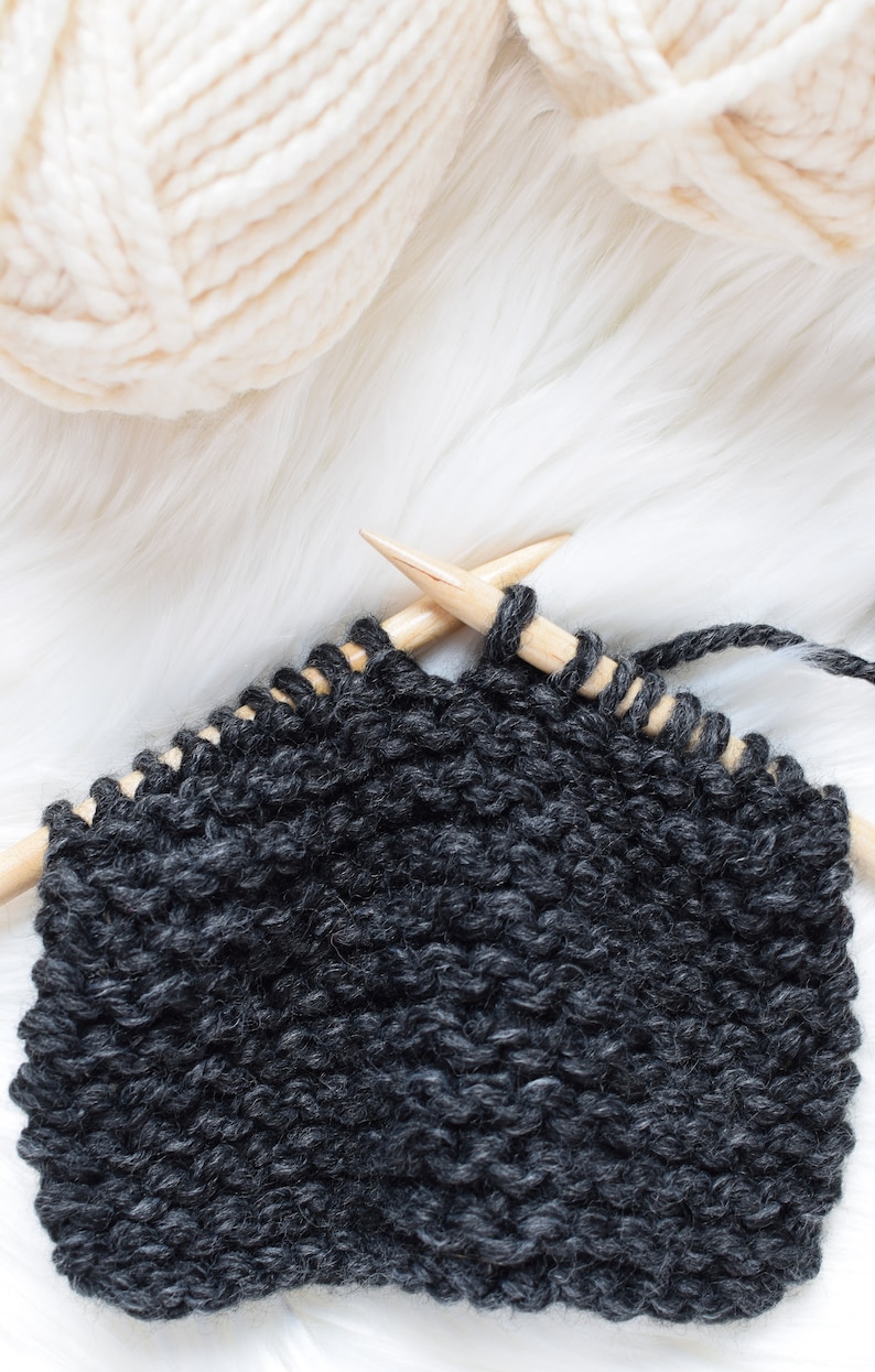 A work in progress photo of a charcoal colored scarf with cream colored yarn in the background.