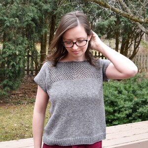 Puede incluir: Un top de punto gris con un patr&oacute;n de encaje alrededor del escote y mangas cortas. El top se lleva con pantalones rojos.