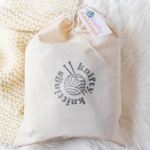 An image of the scarf knitting kit packaged up in a hand stamped project bag, laying on a finished cream colored scarf.