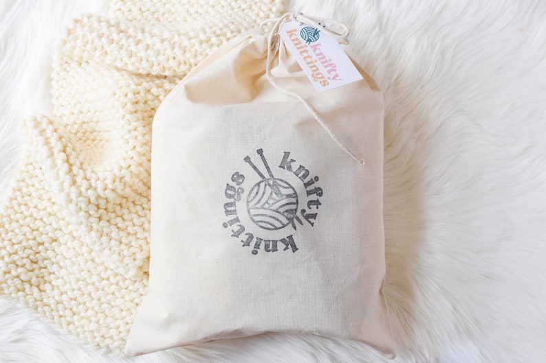 An image of the scarf knitting kit packaged up in a hand stamped project bag, laying on a finished cream colored scarf.