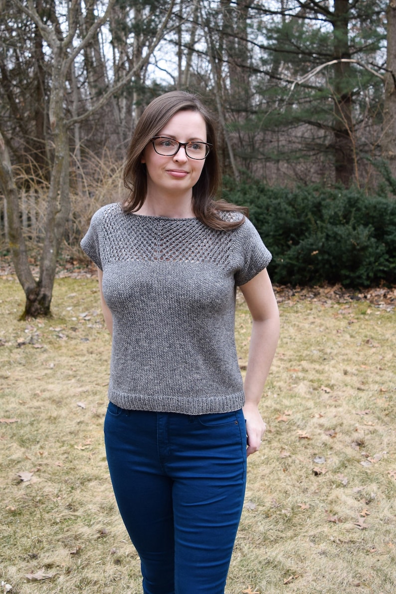Puede incluir: Una mujer lleva una blusa de punto gris con un escote de encaje y unos vaqueros azules. La blusa tiene mangas cortas y un corte holgado.