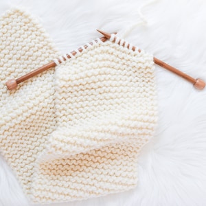 A work in progress photo of a cream colored scarf partially knit and still on the knitting needles.