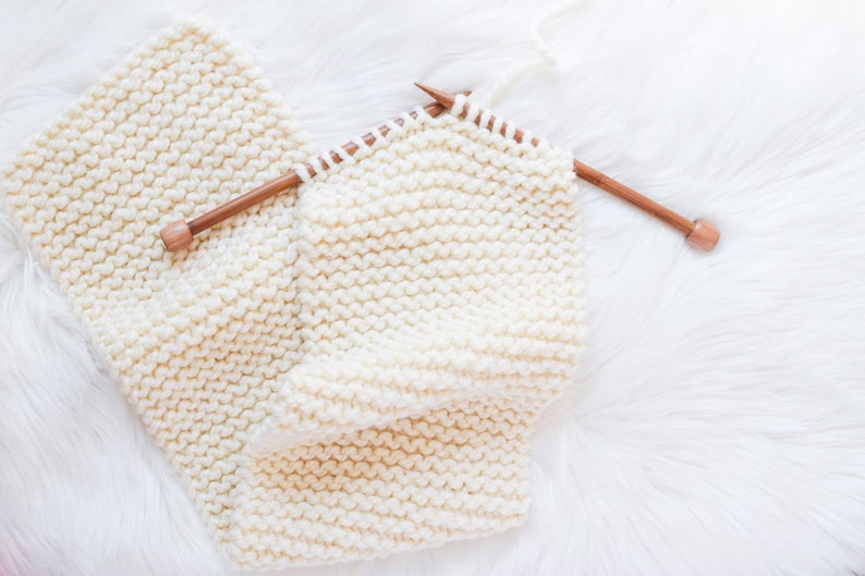 A work in progress photo of a cream colored scarf partially knit and still on the knitting needles.