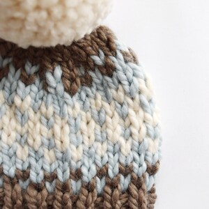 K&ouml;nnte beinhalten: Eine braune, blaue und wei&szlig;e Strickm&uuml;tze mit einem wei&szlig;en Bommel oben. Die M&uuml;tze hat ein braunes Leder-Etikett mit den Buchstaben "KK" darauf.
