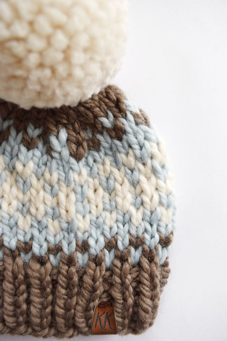 K&ouml;nnte beinhalten: Eine braune, blaue und wei&szlig;e Strickm&uuml;tze mit einem wei&szlig;en Bommel oben. Die M&uuml;tze hat ein braunes Leder-Etikett mit den Buchstaben "KK" darauf.