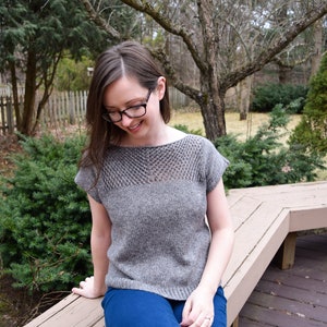 Puede incluir: Una mujer con un top de punto gris con un patr&oacute;n de encaje y jeans azules se sienta en un banco de madera.