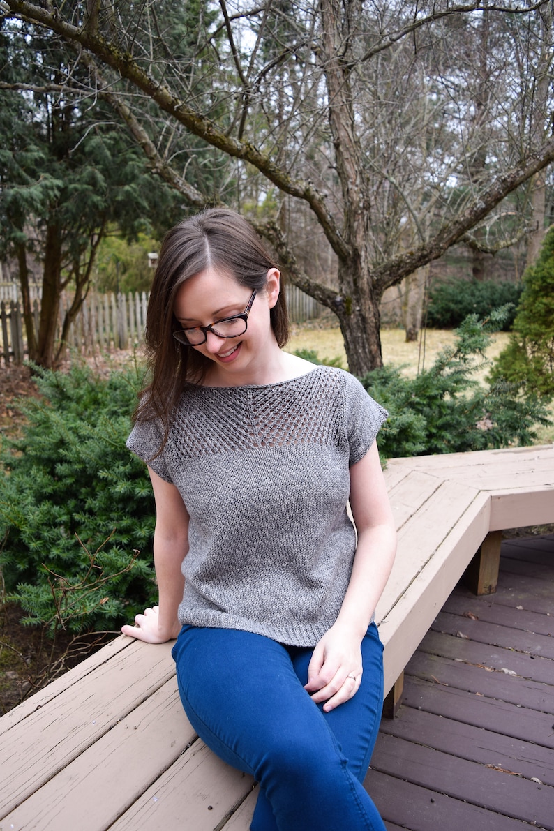 Puede incluir: Una mujer con un top de punto gris con un patr&oacute;n de encaje y jeans azules se sienta en un banco de madera.
