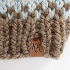 K&ouml;nnte beinhalten: Eine braune Strickm&uuml;tze mit einem blau-wei&szlig; gestreiften Muster. Die M&uuml;tze hat ein braunes Lederlabel mit den Buchstaben "KK" darauf.