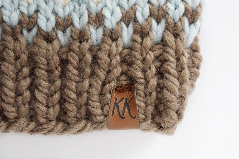 K&ouml;nnte beinhalten: Eine braune Strickm&uuml;tze mit einem blau-wei&szlig; gestreiften Muster. Die M&uuml;tze hat ein braunes Lederlabel mit den Buchstaben "KK" darauf.