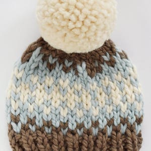 K&ouml;nnte beinhalten: Eine braune, wei&szlig;e und blaue Strickm&uuml;tze mit einem wei&szlig;en Bommel oben. Die M&uuml;tze hat ein braunes Strickband am unteren Rand mit einem braunen Lederanh&auml;nger, auf dem "KK" steht.