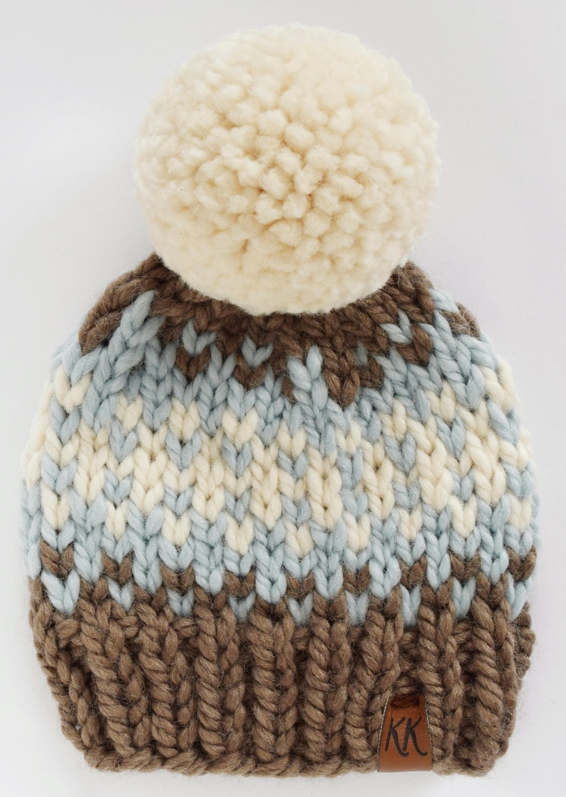 K&ouml;nnte beinhalten: Eine braune, wei&szlig;e und blaue Strickm&uuml;tze mit einem wei&szlig;en Bommel oben. Die M&uuml;tze hat ein braunes Strickband am unteren Rand mit einem braunen Lederanh&auml;nger, auf dem "KK" steht.