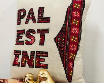 May include: A white pillow with the word "PALESTINE" embroidered in red and a red and black embroidered outline of the country of Palestine. The pillow is sitting on a table with two gold bird figurines in front of it. The text "Digital Download PDF" is in the top right corner of the image. The text "Cross Stitch Pattern ONLY" is in a circle in the bottom right corner of the image. The text "All proceeds will support the Gaza-based artist who created this design" is at the bottom of the image.