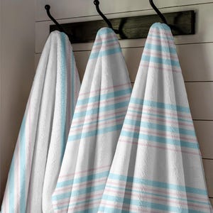 May include: Three white plush towels with blue and pink stripes hang from black hooks on a wooden board. The towels are displayed against a white, slatted wall, creating a clean and inviting aesthetic.