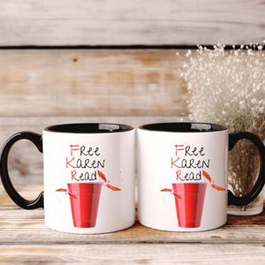 Puede incluir: Dos tazas de café blancas con asas y bordes negros. Cada taza presenta el texto "Free Karen Read" en escritura roja sobre una copa roja con detalles en naranja. Las tazas se exhiben sobre una superficie de madera.