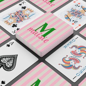 May include: A deck of playing cards featuring a pink and white striped design, with the name "Maisie" printed on the top card. Other cards, including a Joker, King, and Ace of Spades, are visible around the deck.