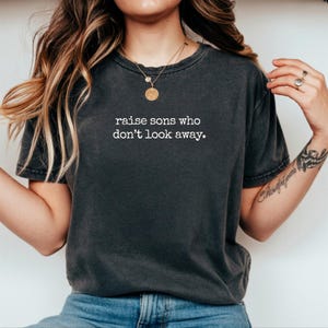 以下が含まれることがあります： 胸元に白い文字で「raise sons who don't look away.」とプリントされたチャコールグレーの半袖Tシャツ。クルーネックで、ヴィンテージ風のウォッシュ加工が施されており、青いデニムジーンズと組み合わされています。