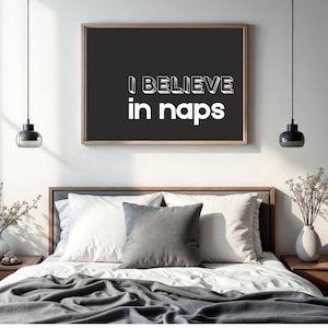 May include: A framed print with the text "I BELIEVE in naps" in white letters on a black background. The print hangs above a bed with white bedding, grey pillows, and a grey blanket. Two pendant lights hang from the ceiling.