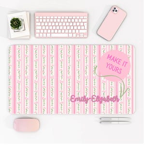 May include: A pink and white striped desk mat with floral accents and the text "Emily Elizabeth". A pink keyboard, mouse, and phone are also visible. The mat has the text "MAKE IT YOURS" in a pink circle.