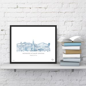 May include: A black framed print of a blue line drawing of the University of North Carolina campus, including the bell tower and the Dean Smith Center. The text "University of North Carolina" and "Chapel Hill, NC" is below the drawing.