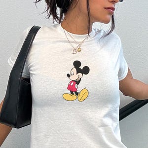 May include: White t-shirt with a Mickey Mouse graphic in a red jacket, yellow shoes, and black shorts. The outfit includes a black bag and a floral skirt. The text "MODEL IS WEARING XSMALL" is visible.
