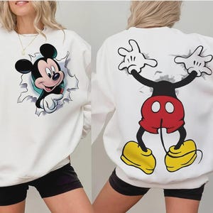 Puede incluir: Sudadera blanca con un diseño colorido de Mickey Mouse en la parte delantera y trasera. El diseño frontal muestra a Mickey rompiendo un efecto de papel rasgado, mientras que la parte trasera muestra el cuerpo y los brazos de Mickey. La sudadera tiene cuello redondo y mangas largas.