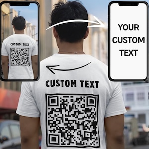 Op de afbeelding: Wit T-shirt met de tekst "CUSTOM TEXT" boven een QR-code op de achterkant. De afbeelding toont een persoon die het shirt draagt, met een smartphone die hetzelfde ontwerp weergeeft. De woorden "YOUR CUSTOM TEXT" staan op het telefoonscherm.