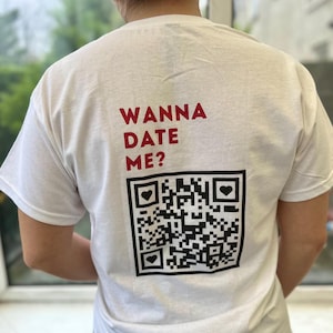 Könnte beinhalten: Weißes T-Shirt mit dem Text "Wanna Date Me?" und einem schwarz-weißen QR-Code mit roten Herzen auf der Rückseite.