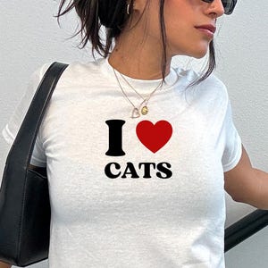 May include: White t-shirt with the text "I ♥ CATS" in black and red. The shirt is a crew neck style. The model is wearing an XSmall size.