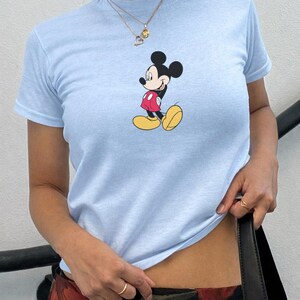 May include: Pale blue t-shirt with a Mickey Mouse cartoon print. The top has short sleeves and a round neck. The cartoon character is shown in his signature red shorts, yellow shoes, and black ears.