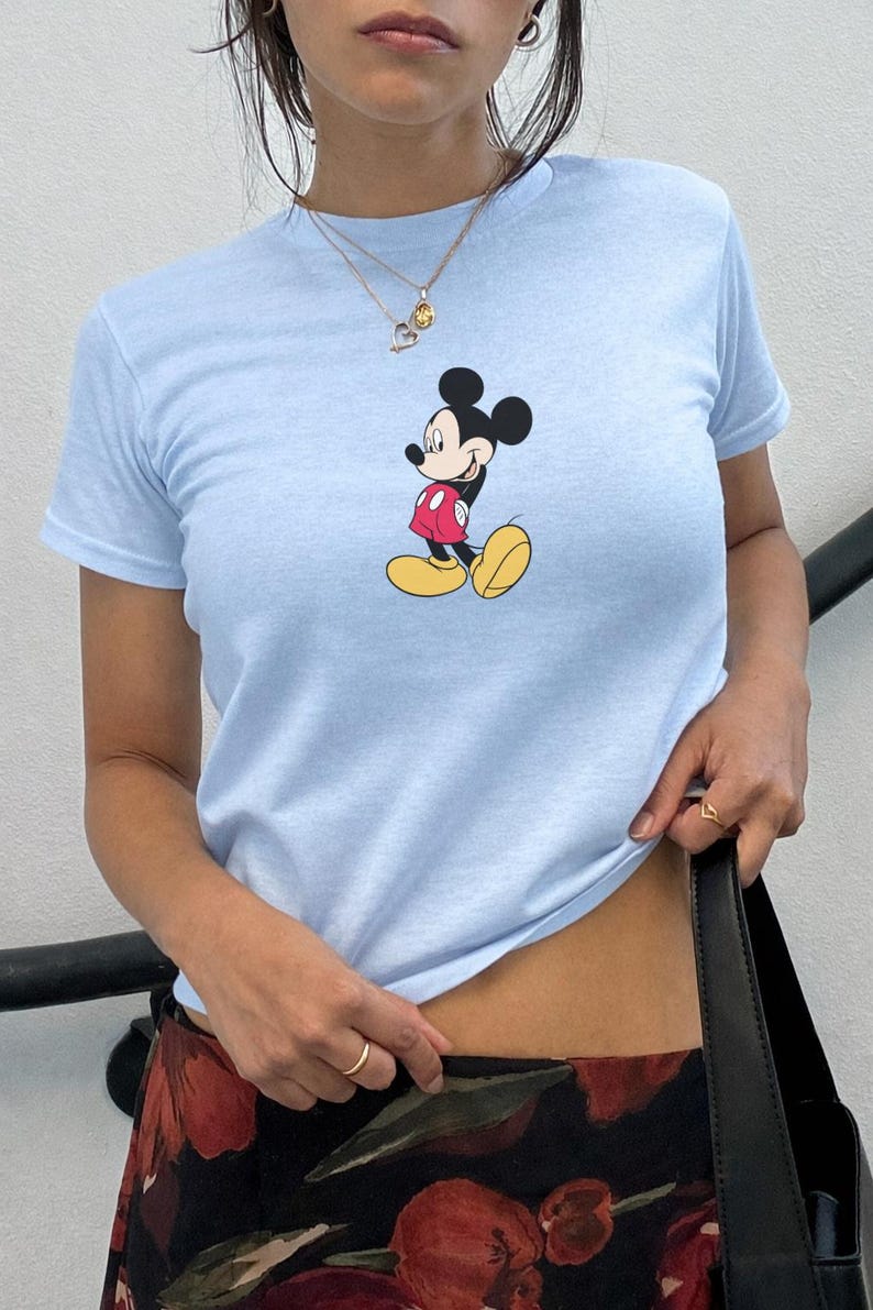May include: Pale blue t-shirt with a Mickey Mouse cartoon print. The top has short sleeves and a round neck. The cartoon character is shown in his signature red shorts, yellow shoes, and black ears.