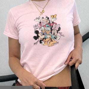 Könnte beinhalten: Hellrosa T-Shirt mit einem farbenfrohen Aufdruck von Disney-Figuren wie Mickey Mouse, Minnie Mouse und Donald Duck. Das Shirt hat einen Rundhalsausschnitt und kurze Ärmel. Der Text "MODEL IS WEARING XSMALL" befindet sich unten.